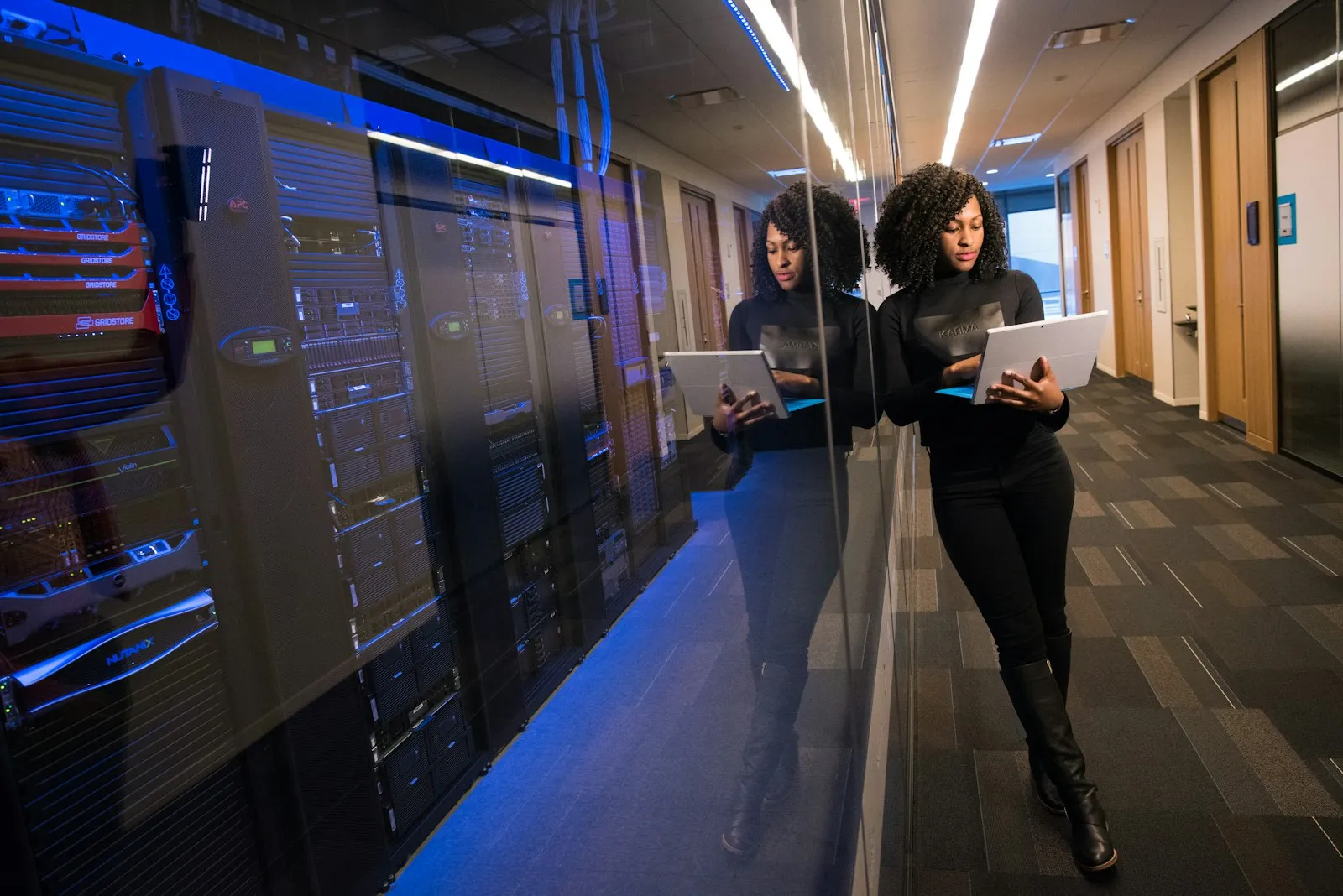 A woman holding an open laptop in an office corridor beside a glass-walled server room